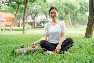 Mutlu, olgun bir kadın parkta bağdaş kurup yoga minderini ve su şişesini işaret eder.