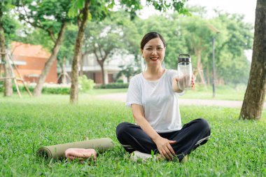 Spor yaptıktan sonra parkta oturan bir kadın elinde su şişesiyle kameraya gülümsüyor.