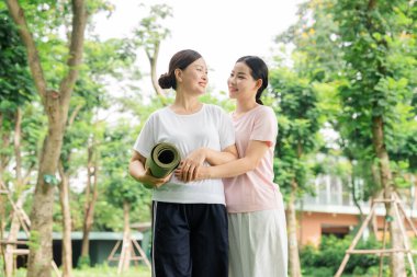 Bir anne ve kızı parkta yoga minderiyle kucaklaşıp spor gününün tadını çıkarıyorlar.