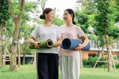 Yeşillik ve ağaçlarla dolu bir ortamda yoga minderleri tutan iki gülen kadın.