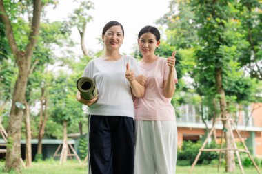 Spor kıyafetli iki neşeli kadın parkta yeşillikle dikiliyorlar. Biri yoga minderi tutuyor.