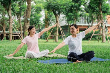 Kadınlar, yeşil çimen ağaçlarıyla çevrili haç bacaklı parkta yoga yapar. Huzurlu bir günün tadını çıkarırlar.