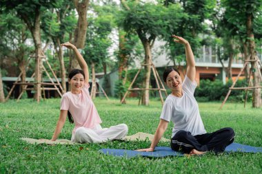 Yoga yapan iki gülümseyen kadın dışarıda, çimenli bir parkta yoga minderlerinin üzerinde oturuyorlar.