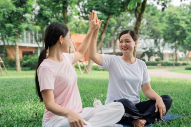 İki gülümseyen kadın, parkta yoga minderinde otururken beşlik çakarak kutluyor.