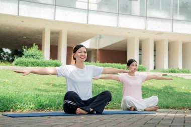 İki kadın, yemyeşil ve modern bir binayla çevrili mavi bir minderde yoga yapıyorlar.
