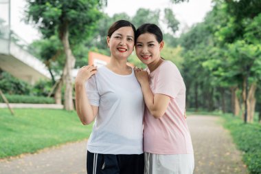 Gülümseyen bir anne ve kız, açık bir park yolunda yemyeşil bir alanda samimi bir kucaklaşmayı paylaşıyor.