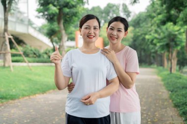 İki kadın parkın dışında, asfalt bir yolda birbirine sarılıyor. Ağaçlar ve yeşil otlarla çevrili.