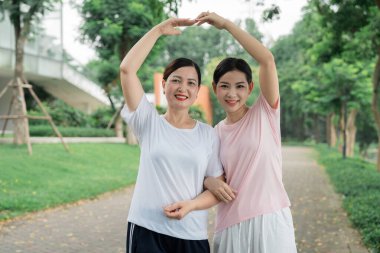 Parkta yan yana duran iki mutlu kadın kollarında gülücüklerle kalp şekli oluşturuyorlar.