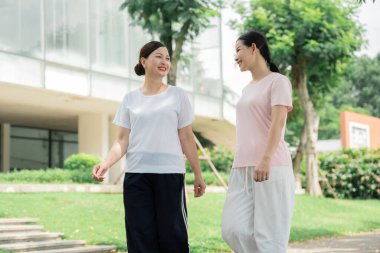 İki gülümseyen kadın parkta yürürler atletik kıyafetler giyerler ve temiz havanın ve sohbetin tadını çıkarırlar.