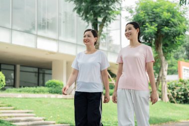 İki gülümseyen kadın, güzel bir günde bir binanın yakınındaki parkta ve ağaçlarda birlikte yürürler.