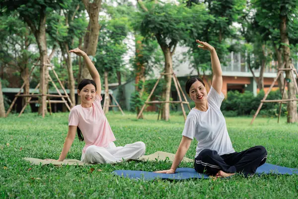 Yoga yapan iki gülümseyen kadın dışarıda, çimenli bir parkta yoga minderlerinin üzerinde oturuyorlar.