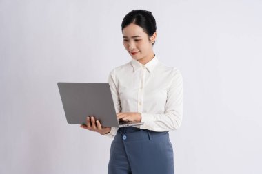 Profesyonel bir kadın gülümsüyor ve minimalist bir ortamda dizüstü bilgisayar kullanıyor. Beyaz gömlek ve pantolon giyiyor.