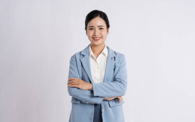 Gülümseyen bir kadın açık mavi bir takım elbise ve beyaz bir bluz giyerek beyaz bir arkaplanda poz verir.