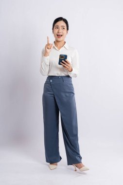 Beyaz bluz ve mavi pantolon giyen bir kadın yukarı doğru işaret ederken akıllı telefon tutuyor.