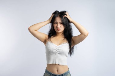 Genç bir kadın kaşları çatık ve gergin görünüyor. Her iki eli de saçlarını çekiyor. Önünde düz beyaz bir arkaplan var.