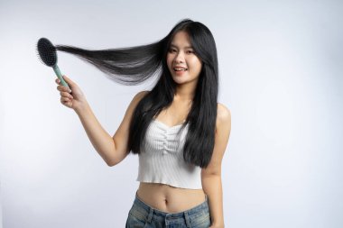 Uzun siyah saçlı, gülümseyen genç bir kadın düz beyaz bir arkaplanda saçını tarıyor.