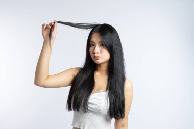 Bir kadın uzun düz siyah saçlarından bir tutam tutuyor Beyaz bir göğüs kafesi giyiyor