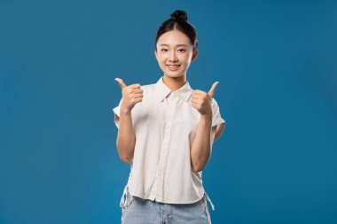 Genç bir kadın gülümsüyor ve mavi arka planda beyaz düğmeli bir gömlek giyerken başparmağını kaldırıyor.