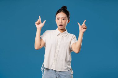 Şaşırmış bir yüz ifadesi olan bir kadın iki eliyle yukarıya doğru işaret eder. Sıradan bir kıyafet giyer.