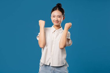 Ekşi saç stili olan genç enerjik bir kadın mavi arka planda gülümseyen yumruklarını kaldırıyor.