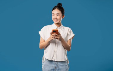 Neşeli bir Asyalı kadın akıllı bir telefon tutuyor ve sol arka planına bakıyor.