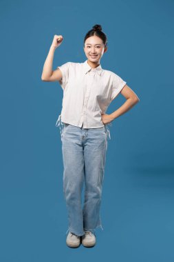 Gülümseyen bir kadın mavi bir arka planda dikilirken güç gösterisi yaparak kolunu esnetiyor.