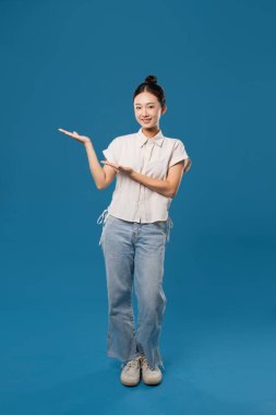 Düğmeli ve kot pantolonlu Asyalı bir kadın açık elle sunum yapıyor. Gülümsüyor.