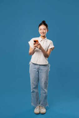 Saf mavi bir arkaplan önünde cep telefonu kullanan bir kadının gülümsemesinin tam görüntüsü.