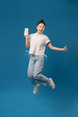 Gülümseyen bir kadın, elinde boş bir cep telefonuyla mavi bir arka planın önüne atlarken görülüyor.