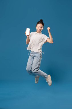 Genç bir kadın elinde bir cep telefonuyla sevinçten havalara uçuyor.