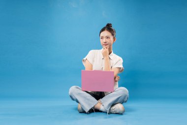 Maviye bürünmüş genç bir kadın dizüstü bilgisayarla bağdaş kurup dalgın dalgın bakıyor.