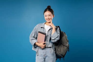 Sırt çantası ve kitapla gülümseyen genç bir kadın mavi bir arka plana yaslanmış düşünceli görünüyor.