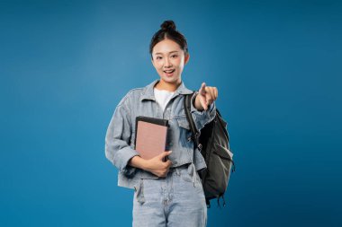 Sırt çantası ve kitapları olan arkadaş canlısı bir kadın mavi arka plandaki izleyiciyi işaret ediyor.