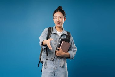 Kitapları ve sırt çantasıyla neşeli bir bayan öğrenci mavi bir arka plana karşı bir karşılama eli uzatıyor.