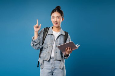 Kot ceketli, sırt çantalı ve kitaplı gülümseyen genç bir kadın mavi stüdyo arka planına işaret ediyor.