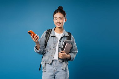 Elinde kitap, elinde telefon ve sırt çantası olan bir öğrenci okula gitmek için mavi arka planda hazır.