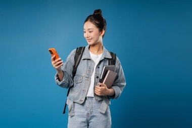 Kitapları ve sırt çantası olan genç bir kadın mavi arka plan eğitimi ve teknoloji konsepti için telefon kullanıyor.