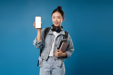 Sırt çantası ve kitaplarıyla gülümseyen öğrenci öğrenmeye hevesli mavi bir arka planda telefon ekranı sergiliyor.