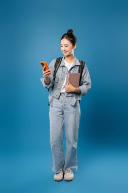 Gündelik kıyafetli genç bir kadın, canlı arkaplana karşı telefon kullanırken bir kitap ve sırt çantası taşıyor.