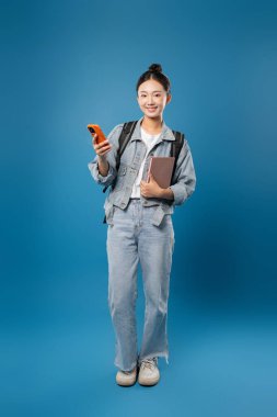 Sırt çantası ve telefonuyla gülümseyen bir öğrenci. Parlak mavi bir arka planda günlük kot pantolon giymiş.