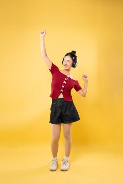 Neşeli bir yüzü olan bir kadın, parlak sarı bir fon karşısında, kulaklıklarıyla enerjik bir şekilde dans eder.