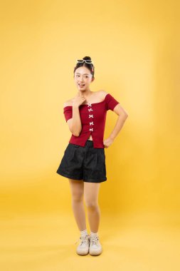 Sarı arka planda poz veren genç bir kadın koyu kırmızı şort ve beyaz spor ayakkabı giyiyor.