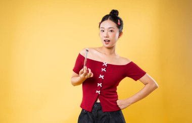 Genç bir bayan elinde makyaj fırçası, sarı bir zeminde sürpriz bir ifadeyle poz veriyor.