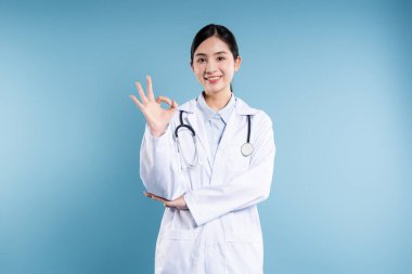 Beyaz doktor önlüğü ve steteskop giymiş gülümseyen bir kadın mavi arka planda iyi bir el hareketi yapar.