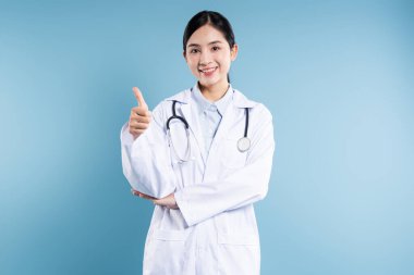 Beyaz önlüklü gülümseyen Asyalı bir doktor mavi arka plan önünde başparmak kaldırıyor.