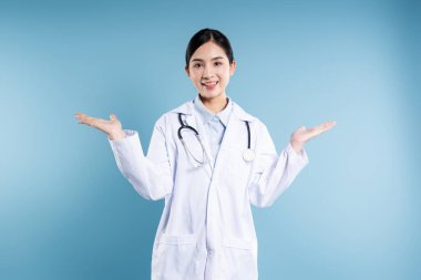 Beyaz önlüklü genç bir doktor avuçlarını açık bir şekilde mavi arka planda sıcak bir gülümseme ile sunar.