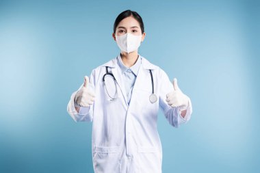 Laboratuar önlüklü, steteskop maskeli ve eldivenli profesyonel doktor mavi arka planda baş parmak hareketi yapıyor.