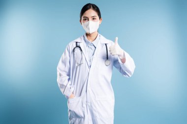 Beyaz önlüklü bir doktor mavi arka planda maske ve steteskop takarak onay veriyor.