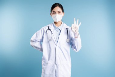 Steteskop maskeli Asyalı tıp uzmanı. Tamam işareti veriyor. Mavi zemin.