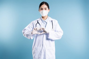 Beyaz önlüklü ve maskeli bir kadın, elleri mavi bir zeminde kalp şekli oluşturur.